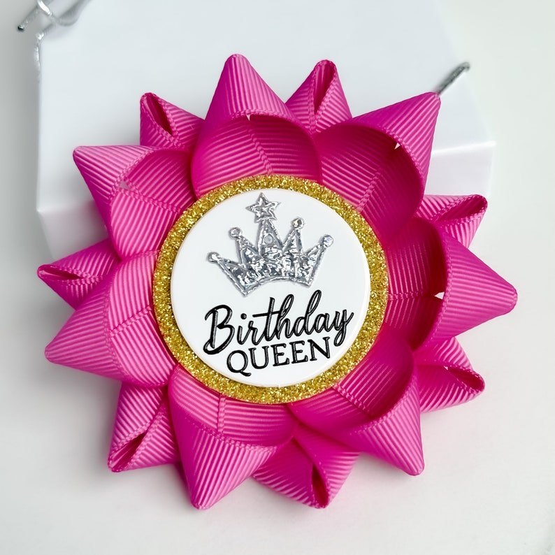 May include: A pink fabric bow with a white circle in the center. The circle has a silver crown with a star on top and the text "Birthday QUEEN" in black.
