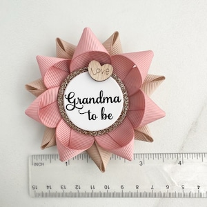 May include: A pink and beige bow with a white circle that says "Grandma to be" and a small wooden heart that says "Love".