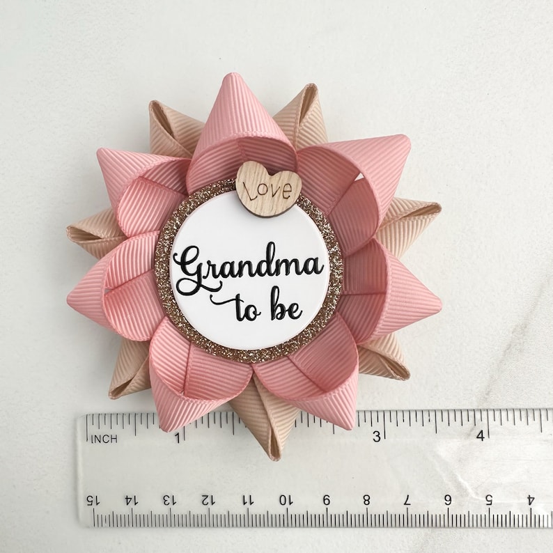 May include: A pink and beige bow with a white circle that says "Grandma to be" and a small wooden heart that says "Love".