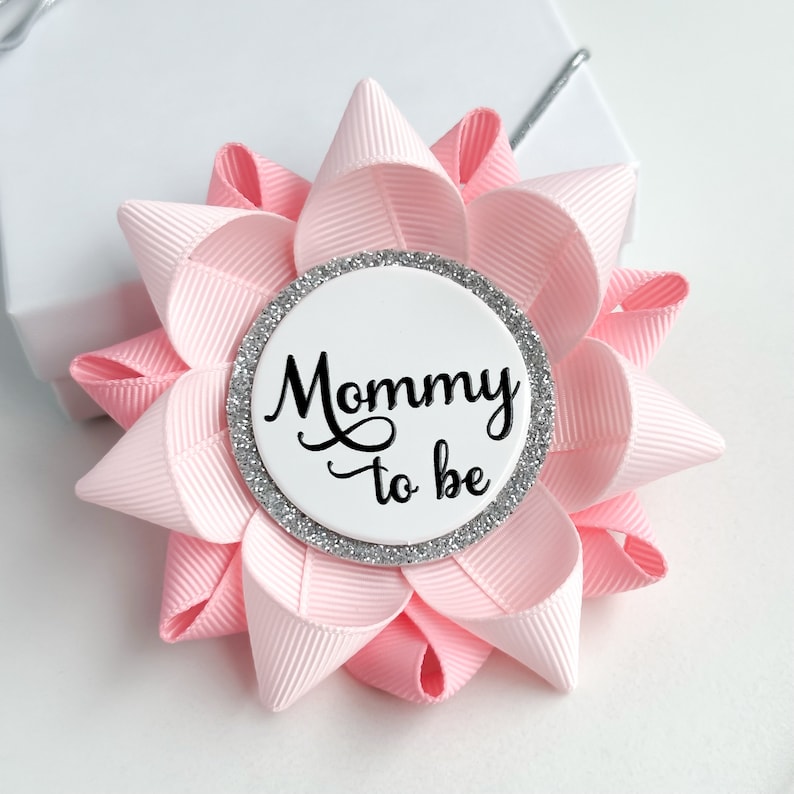 May include: Pink and white flower-shaped button with a silver glitter circle and the text "Mommy to be" in black.