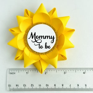 K&ouml;nnte beinhalten: Gelbe, bl&uuml;tenf&ouml;rmige Schleife aus Stoff mit einem wei&szlig;en Kreis in der Mitte, auf dem in schwarzer Schrift "Mommy to be" steht. Der Kreis hat einen goldenen Glitzerrand.