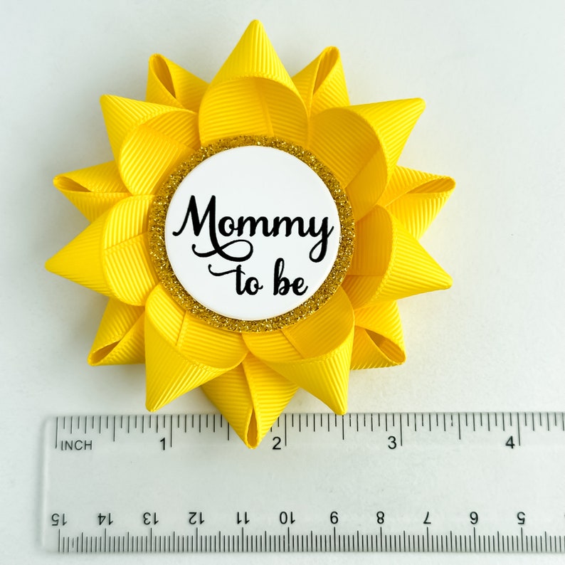 K&ouml;nnte beinhalten: Gelbe, bl&uuml;tenf&ouml;rmige Schleife aus Stoff mit einem wei&szlig;en Kreis in der Mitte, auf dem in schwarzer Schrift "Mommy to be" steht. Der Kreis hat einen goldenen Glitzerrand.