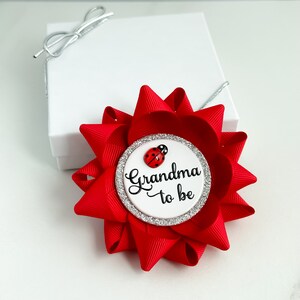 May include: Red and white grandma to be pin with a ladybug. The pin is decorated with silver glitter.