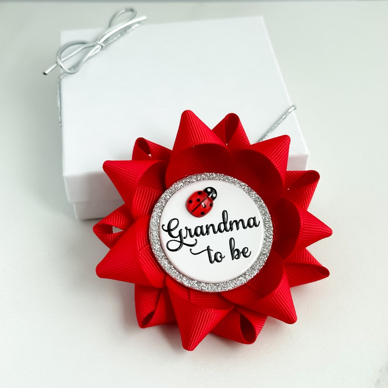 May include: Red and white grandma to be pin with a ladybug. The pin is decorated with silver glitter.