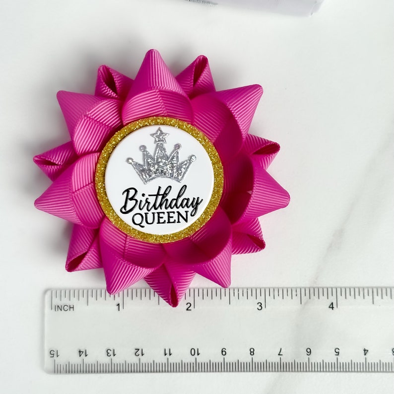 May include: A pink bow with a white circle in the center. The circle has a gold glitter border and a silver crown with a star on top. The text "Birthday Queen" is printed in black inside the circle.