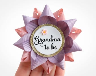 Könnte beinhalten: Blumenförmige Anstecknadel mit einem weißen Mittelteil, auf dem in schwarzer Schrift "Grandma to be" steht. Die Anstecknadel besteht aus rosa und lavendelfarbenen Bändern mit einem goldglitzernden Rand.