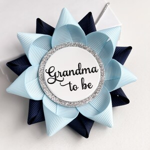 May include: A blue and navy blue bow with a white circle in the centre that says "Grandma to be" in black script. The circle has a silver glitter border.