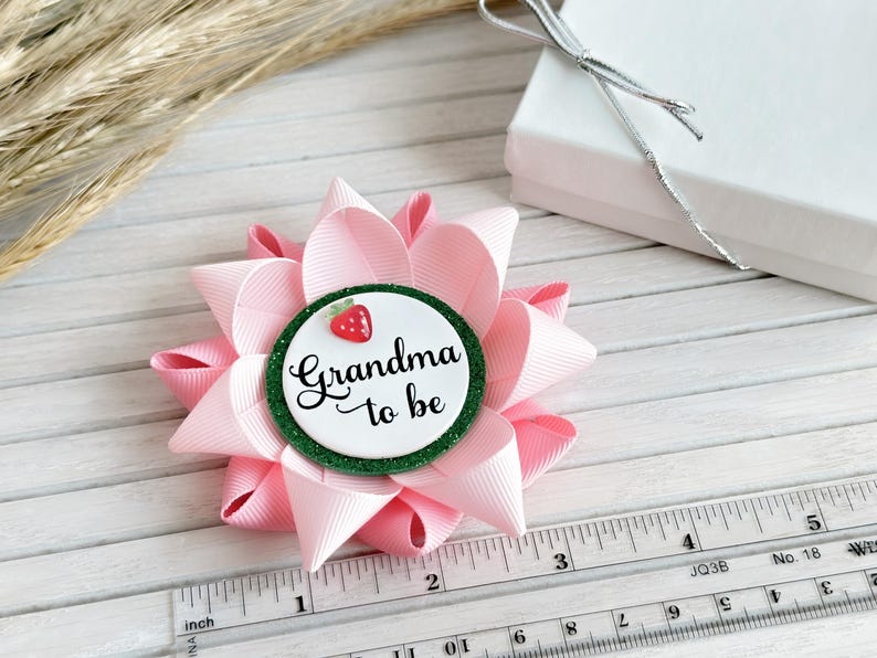May include: Pink and green bow with a white circle that says "Grandma to be" and a red strawberry on top. The bow is about 3 inches in diameter.
