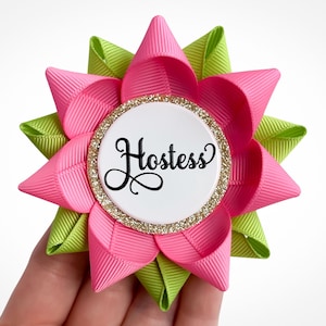 May include: A pink and green fabric flower with a white circle in the center that says "Hostess". The circle has a gold glitter border.
