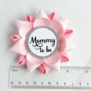 May include: Pink and white flower-shaped pin with a silver glitter circle in the center. The circle has the text "Mommy to be" in black.
