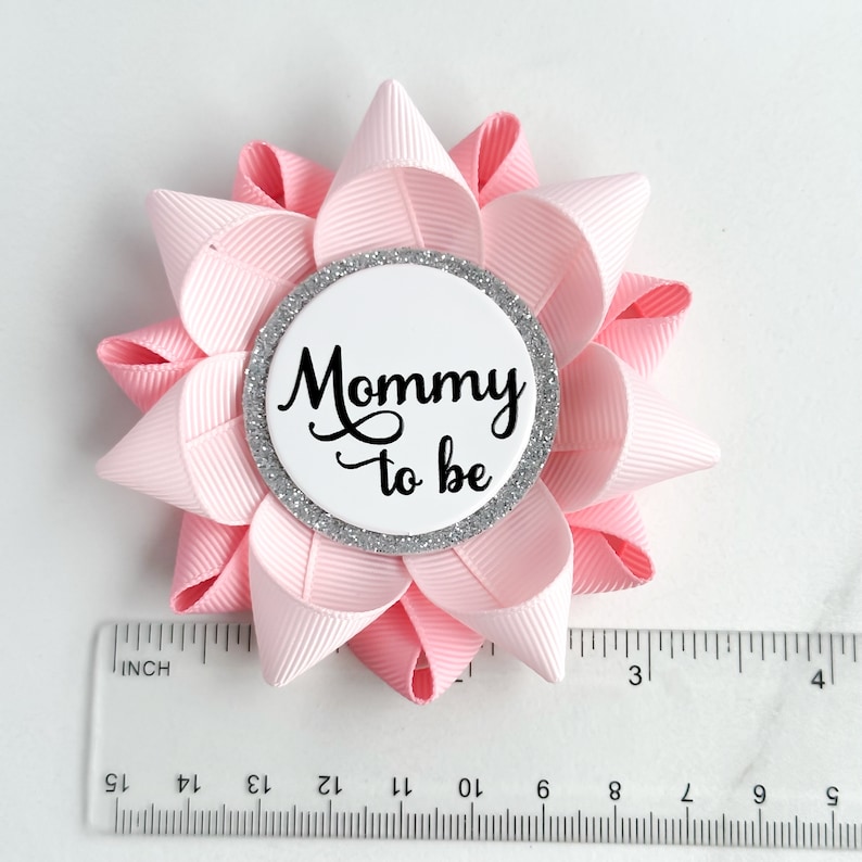 May include: Pink and white flower-shaped pin with a silver glitter circle in the center. The circle has the text "Mommy to be" in black.