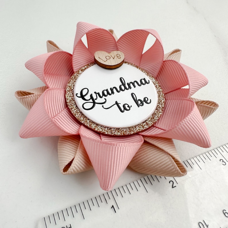 May include: Pink and beige bow with a white circle that says "Grandma to be" and a small wooden heart that says "Love".