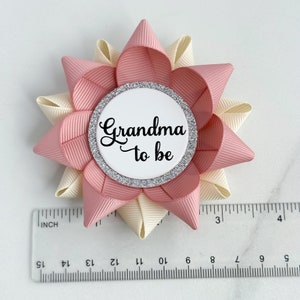 May include: A pink and ivory bow with a silver glitter circle that says "Grandma to be" in black script.