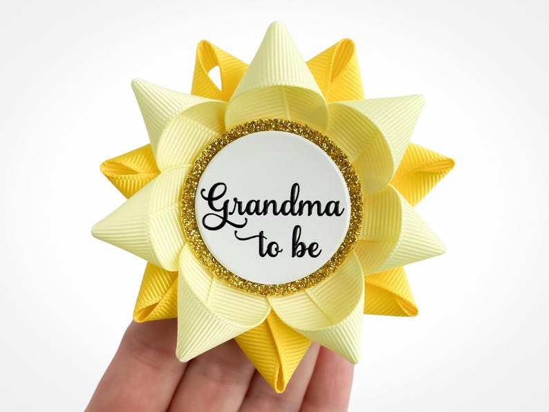 Baby shower pin made of light yellow and yellow ribbon in the shape of a flower with gold accent and custom text: Grandma to be