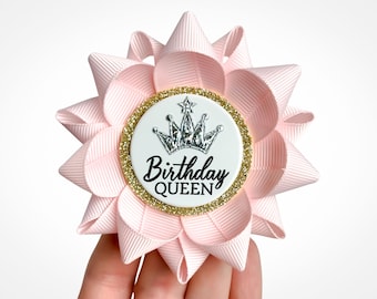May include: Pink fabric flower with a white circle in the center. The circle has a gold glitter border and a silver crown with a star on top. The text "Birthday QUEEN" is printed in black inside the circle.
