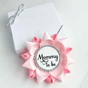 May include: Pink and silver mommy-to-be button with a white background and the text "Mommy to be" in black cursive lettering.