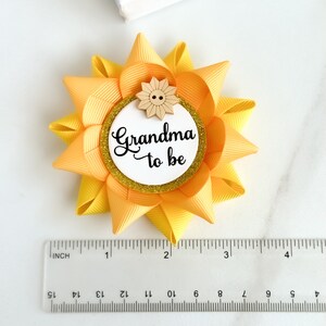 May include: Yellow and orange sunflower-shaped bow with a white circle in the center that says "Grandma to be" and a small wooden sunflower button.