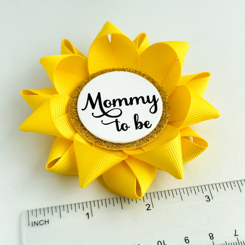 K&ouml;nnte beinhalten: Gelbe bl&uuml;tenf&ouml;rmige Schleife mit einem wei&szlig;en Kreis in der Mitte, auf dem in schwarzer Schrift "Mommy to be" steht. Der Kreis hat einen goldenen Glitzerrand.
