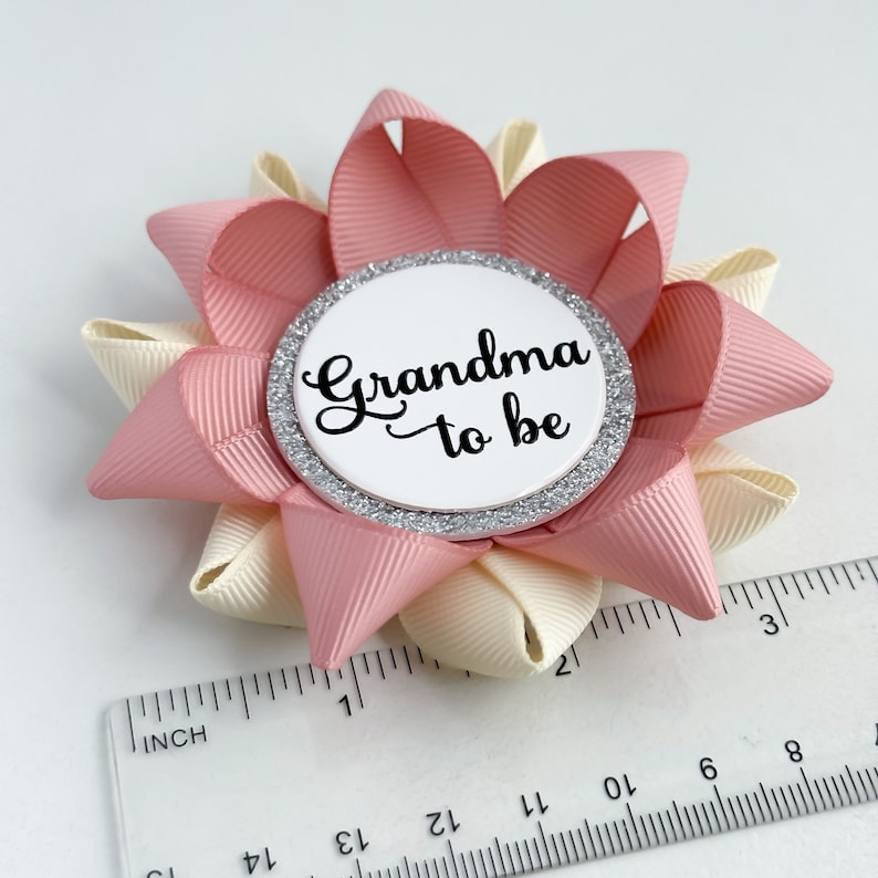May include: A pink and cream flower-shaped button with a silver glitter circle that says "Grandma to be" in black text.