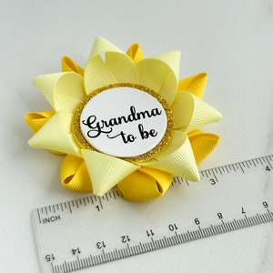 K&ouml;nnte beinhalten: Eine gelbe und hellgelbe Schleife mit einem wei&szlig;en Kreis in der Mitte, auf dem in schwarzer Schrift "Grandma to be" steht. Die Schleife hat einen Durchmesser von etwa 7,5 cm.