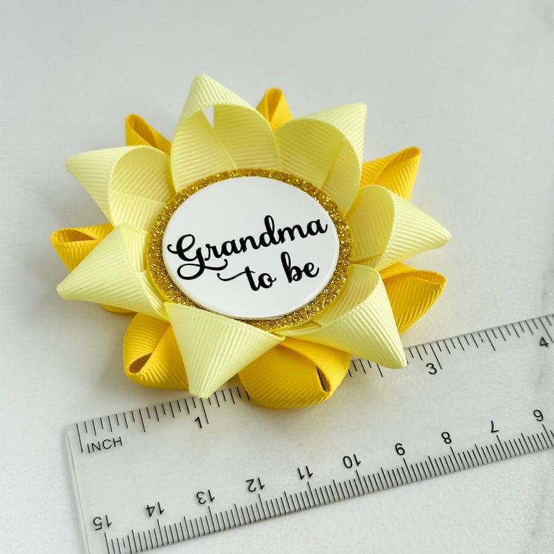 K&ouml;nnte beinhalten: Eine gelbe und hellgelbe Schleife mit einem wei&szlig;en Kreis in der Mitte, auf dem in schwarzer Schrift "Grandma to be" steht. Die Schleife hat einen Durchmesser von etwa 7,5 cm.