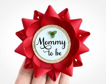 May include: A red ribbon flower with a white central disc. The disc features the words "Mommy to be" in black script, a gold glitter border, and a small holly sprig. A festive accessory.