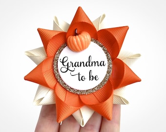 Op de afbeelding: Oranje en witte stoffen bloem met een pompoen en de tekst "Oma in spe" op een witte cirkel met gouden glitter.