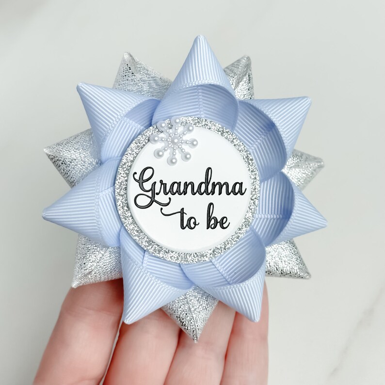 K&ouml;nnte beinhalten: Eine hellblaue und silberne Schleife mit einem wei&szlig;en Kreis in der Mitte. Der Kreis hat einen silbernen Glitzerrand und ein Schneeflockenmuster. Der Text "Grandma to be" ist in Schwarz auf dem Kreis geschrieben.