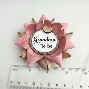 May include: Pink and beige bow with a white circle in the center that says "Grandma to be" with a small wooden heart that says "Love" on top. The bow is about 2 inches in diameter.