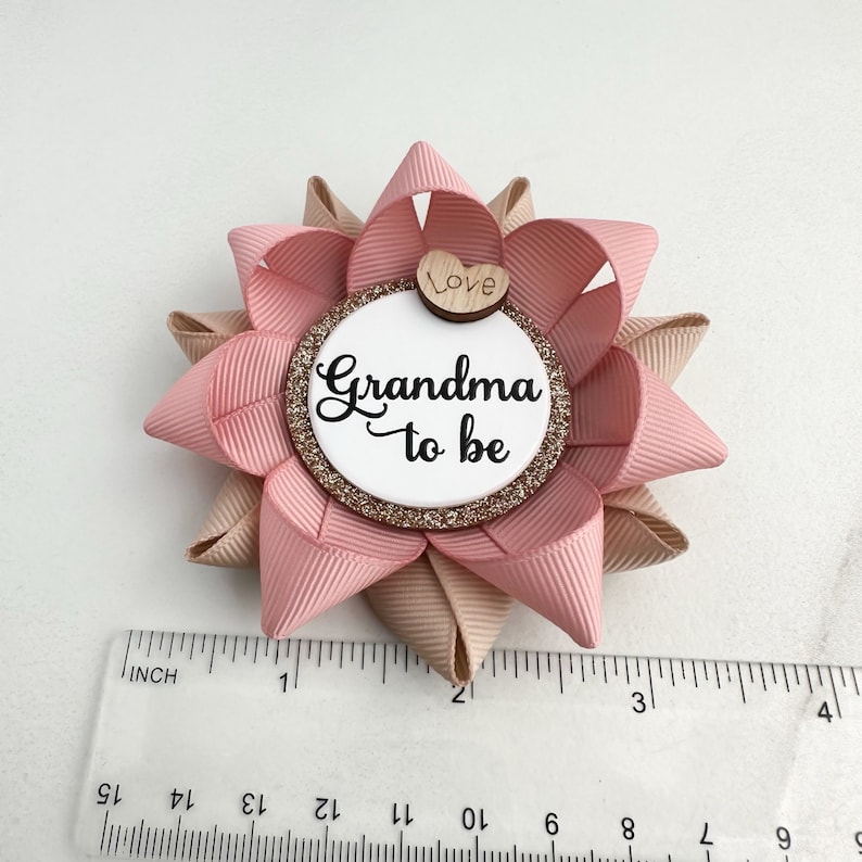 May include: Pink and beige bow with a white circle in the center that says "Grandma to be" with a small wooden heart that says "Love" on top. The bow is about 2 inches in diameter.