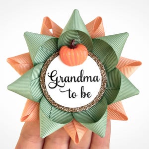 May include: A green and peach bow with a white circle in the center. The circle has gold glitter around the edge and the text "Grandma to be" in black. A small orange pumpkin is on top of the circle.