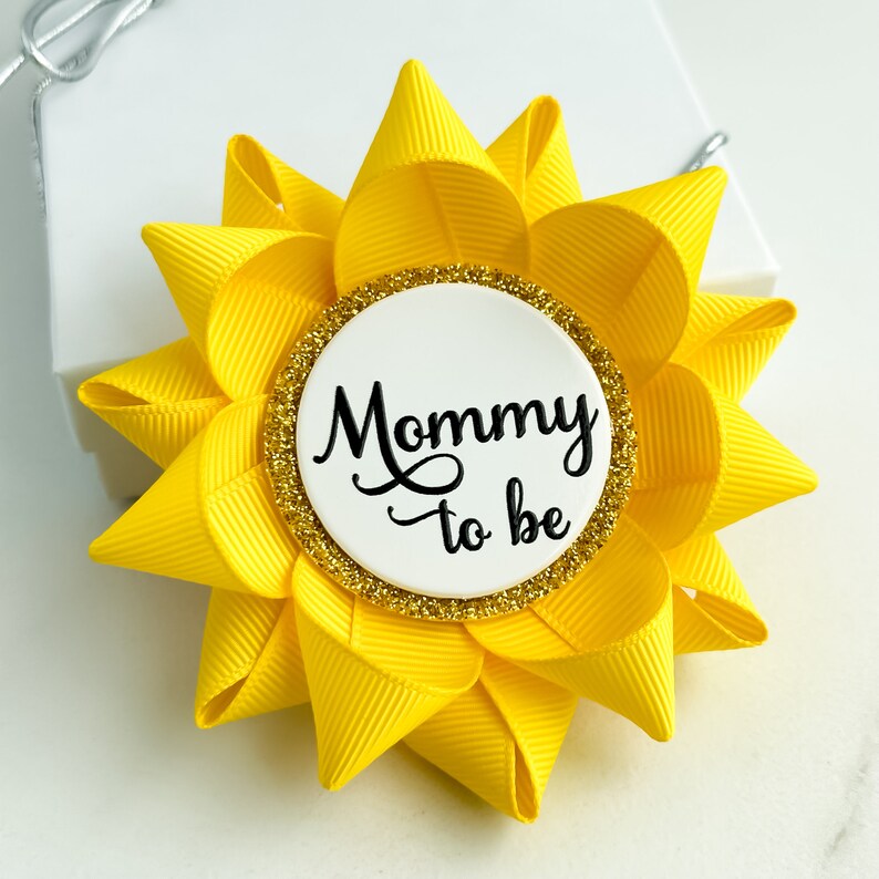 K&ouml;nnte beinhalten: Gelbe, blumenf&ouml;rmige Anstecknadel mit einem wei&szlig;en Kreis in der Mitte, auf dem in schwarzer Schrift "Mommy to be" steht. Der Kreis hat einen goldenen Glitzerrand.