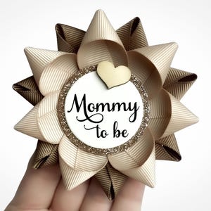 May include: A beige and brown bow with a white circle in the center. The circle has gold glitter around the edge and the text "Mommy to be" in black. A small wooden heart is above the text.