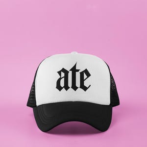 May include: A black and white trucker hat with the word "ate" in a gothic font across the front. The hat has a white front panel, a black brim, and a black mesh back. The background is a solid pink.