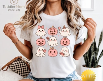 May include: White t-shirt featuring a repeating pattern of cute ghosts and pink pumpkins. The ghosts have bows, flowers, and candy. The text "Toddler Mom DESIGN" is at the top left.