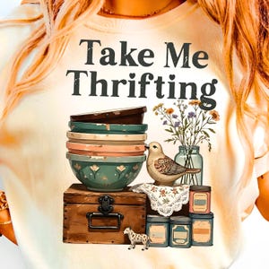 May include: Cream-colored t-shirt with the text "Take Me Thrifting." The graphic features a stack of bowls, a bird, flowers, a wooden chest, and small jars. The design has a vintage, rustic aesthetic.