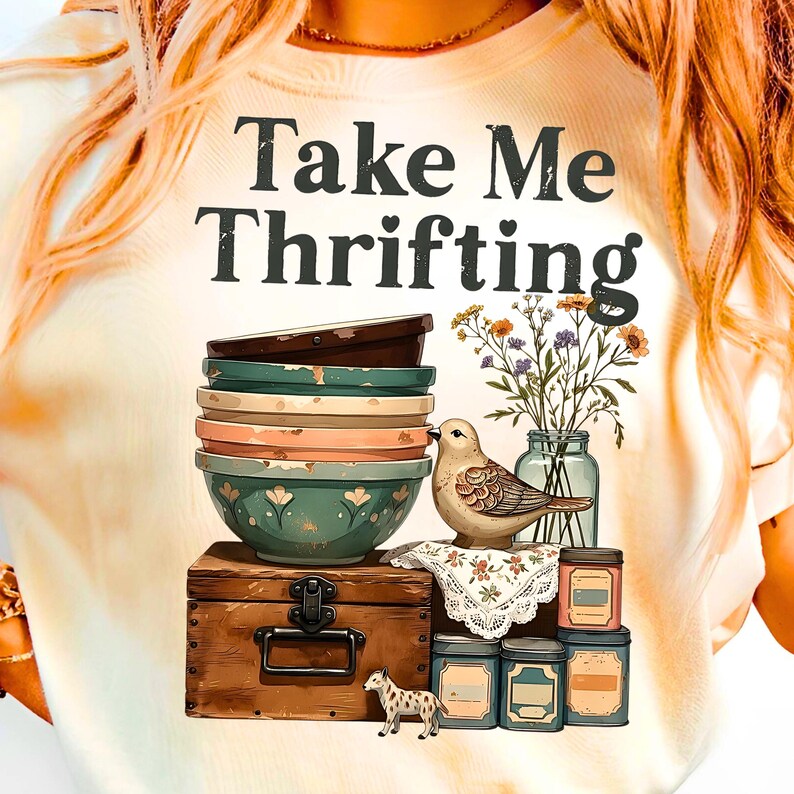 May include: Cream-colored t-shirt with the text "Take Me Thrifting." The graphic features a stack of bowls, a bird, flowers, a wooden chest, and small jars. The design has a vintage, rustic aesthetic.