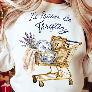 Peut inclure: Sweat-shirt blanc avec l'inscription "I'd Rather Be Thrifting". Un chariot de courses doré contient de la porcelaine bleue et blanche, des cadres et de la lavande. Un tissu rose et beige est également dans le chariot.