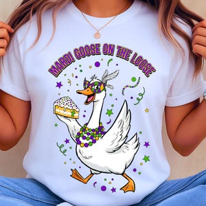 May include: White t-shirt with a cartoon goose wearing a Mardi Gras mask and holding a slice of cake. The text "MARDI GOOSE ON THE LOOSE" is arched above the goose in purple and green. The design includes confetti and stars.