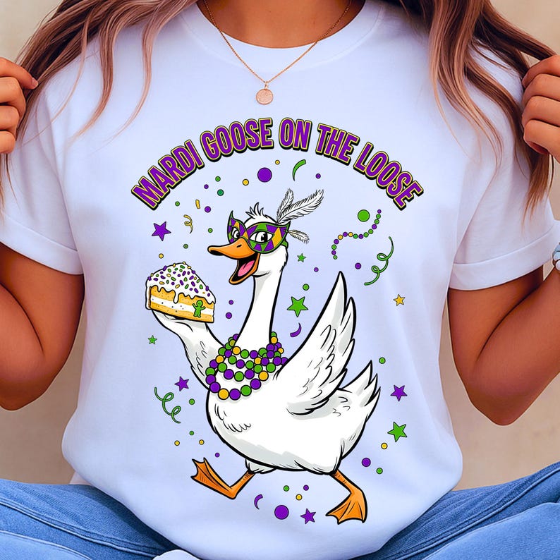 May include: White t-shirt with a cartoon goose wearing a Mardi Gras mask and holding a slice of cake. The text "MARDI GOOSE ON THE LOOSE" is arched above the goose in purple and green. The design includes confetti and stars.