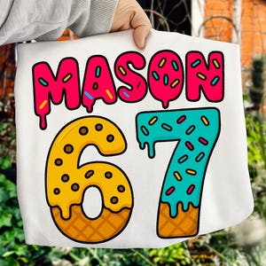 May include: A white fabric item with the name "MASON" in pink, dripping letters, and the number "67" in a donut-style design. The "6" is yellow with brown sprinkles, and the "7" is teal with colorful sprinkles.