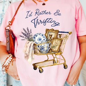 May include: Pink t-shirt with the text "I'd Rather Be Thrifting" in navy blue script. The graphic features a gold shopping cart filled with vintage items, including a blue and white china set, framed pictures, and lavender sprigs.
