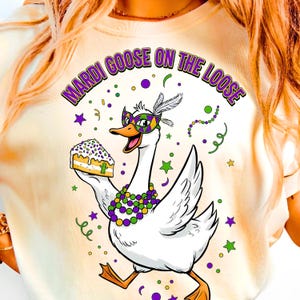 May include: Cream-colored t-shirt featuring a cartoon goose wearing a Mardi Gras mask and holding a slice of cake. The text "MARDI GOOSE ON THE LOOSE" arches above the goose in purple and green. The design includes stars and confetti.