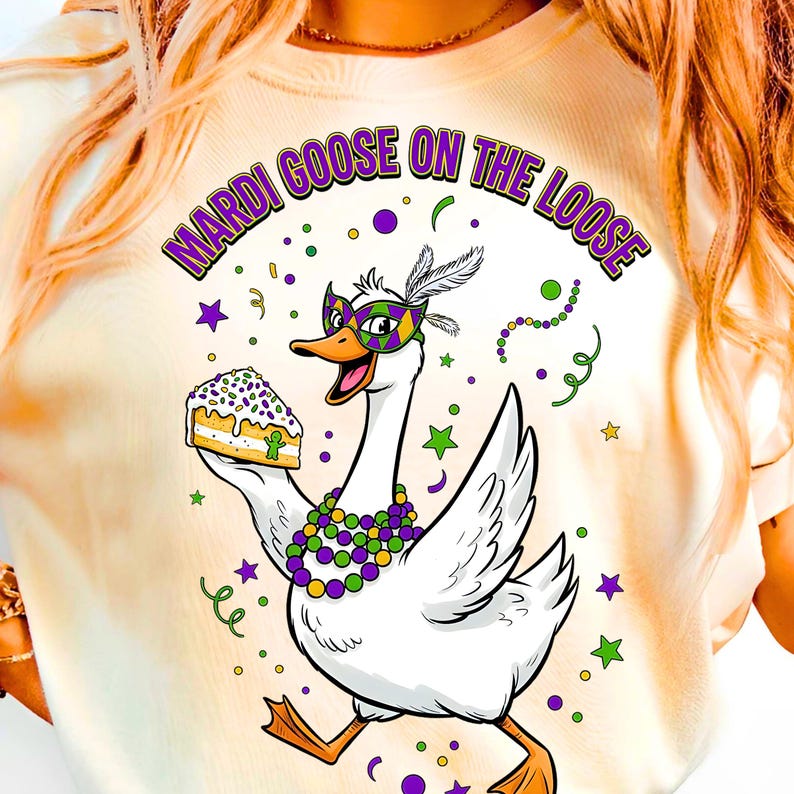 May include: Cream-colored t-shirt featuring a cartoon goose wearing a Mardi Gras mask and holding a slice of cake. The text "MARDI GOOSE ON THE LOOSE" arches above the goose in purple and green. The design includes stars and confetti.