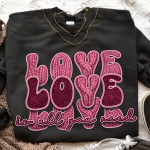 Pode incluir: Camisola preta com as palavras "LOVE LOVE LOVE" em letras rosa brilhantes. A palavra "LOVE" também está num design de malha. A frase "is all you need" está escrita abaixo em script rosa.