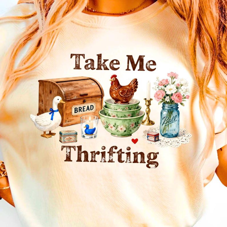 May include: Cream-colored t-shirt with the words "Take Me Thrifting" in brown lettering. The design includes a wooden bread box, a duck with a blue bow, a chicken, bowls, a candle, and a jar of flowers.