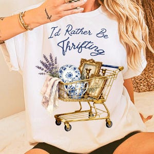 May include: White t-shirt with the text "I'd Rather Be Thrifting" in blue script. The shirt features a gold shopping cart filled with blue and white china, framed pictures, and lavender sprigs.