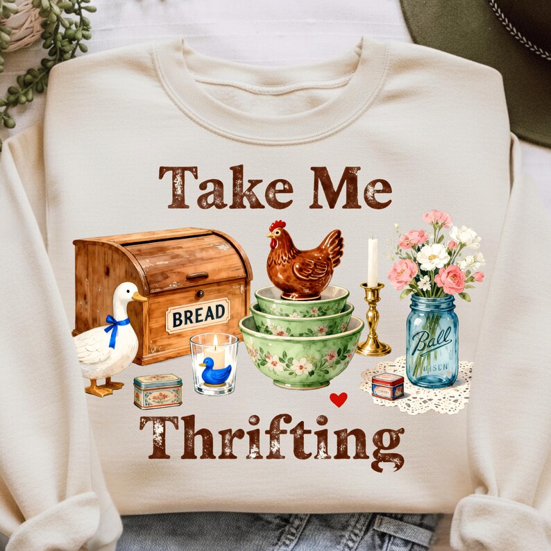 May include: Cream-colored sweatshirt with the text "Take Me Thrifting" in brown. The design includes a wooden bread box, a duck, a chicken, floral bowls, a candle, and a jar of flowers.