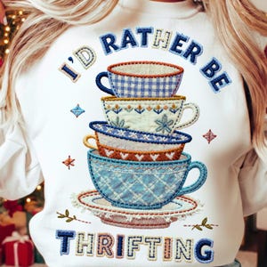 May include: White sweatshirt with embroidered text and stacked teacups. The text reads "I'd Rather Be Thrifting" in blue and tan. The teacups are blue, white, and orange with various patterns. The design includes decorative floral and geometric elements.