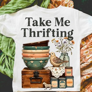 May include: White t-shirt with the text "Take Me Thrifting." The graphic features a stack of bowls, a bird, flowers in a jar, and a wooden chest. Additional elements include small jars and a spotted animal figurine.